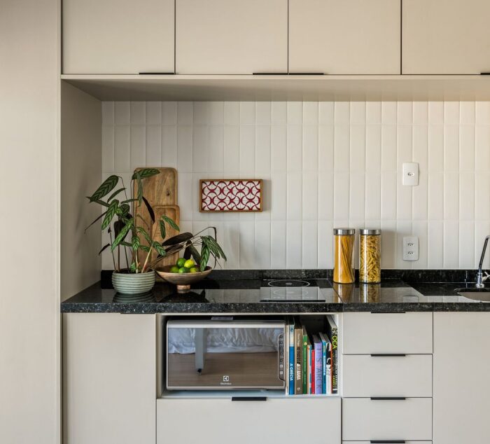 O uso de cores claras nas paredes e móveis contribui para ampliar visualmente a cozinha compacta, criando um ambiente mais arejado e acolhedor. Cozinha com armários claros e objetos bem organizados na bancada.