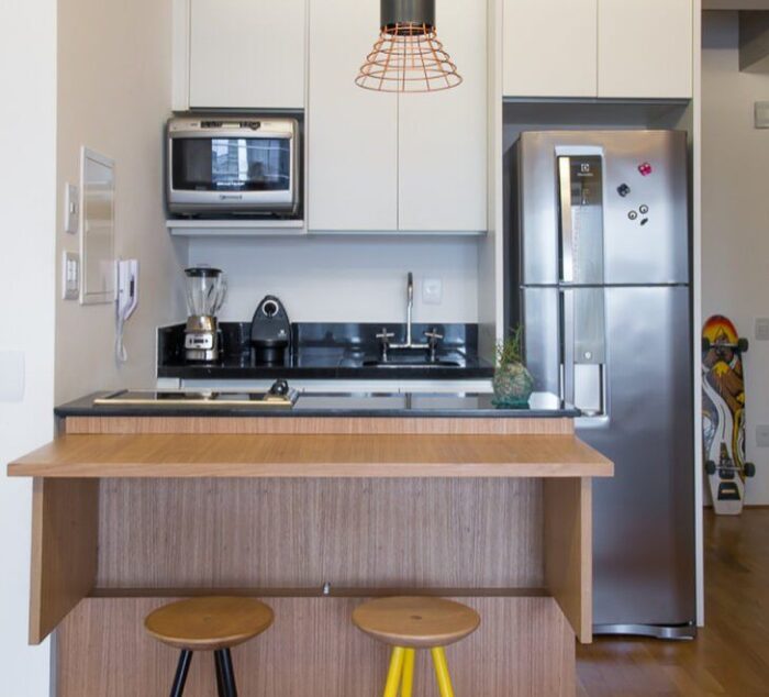 O uso de uma bancada americana integrada à cozinha permite refeições rápidas e ainda economiza espaço, uma escolha perfeita para apartamentos menores. Cozinha pequena com bancada de madeira e banquetas.