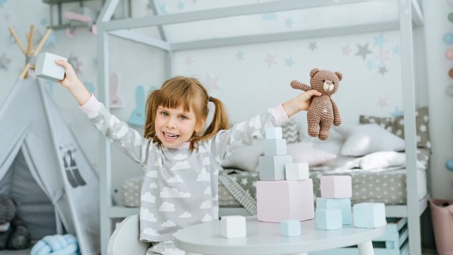 Menina brincando em um quarto infantil decorado com cores suaves, brinquedos e cama estilo cabana ao fundo.