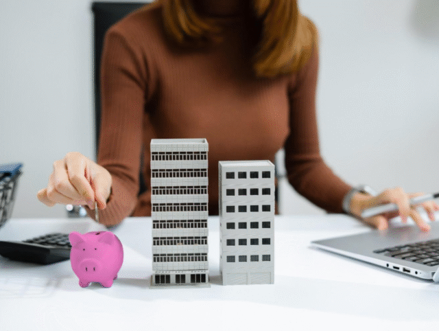 Mulher coloca moeda em cofre ao lado de maquetes de prédios, simbolizando investimento em imóveis financiados para alugar.